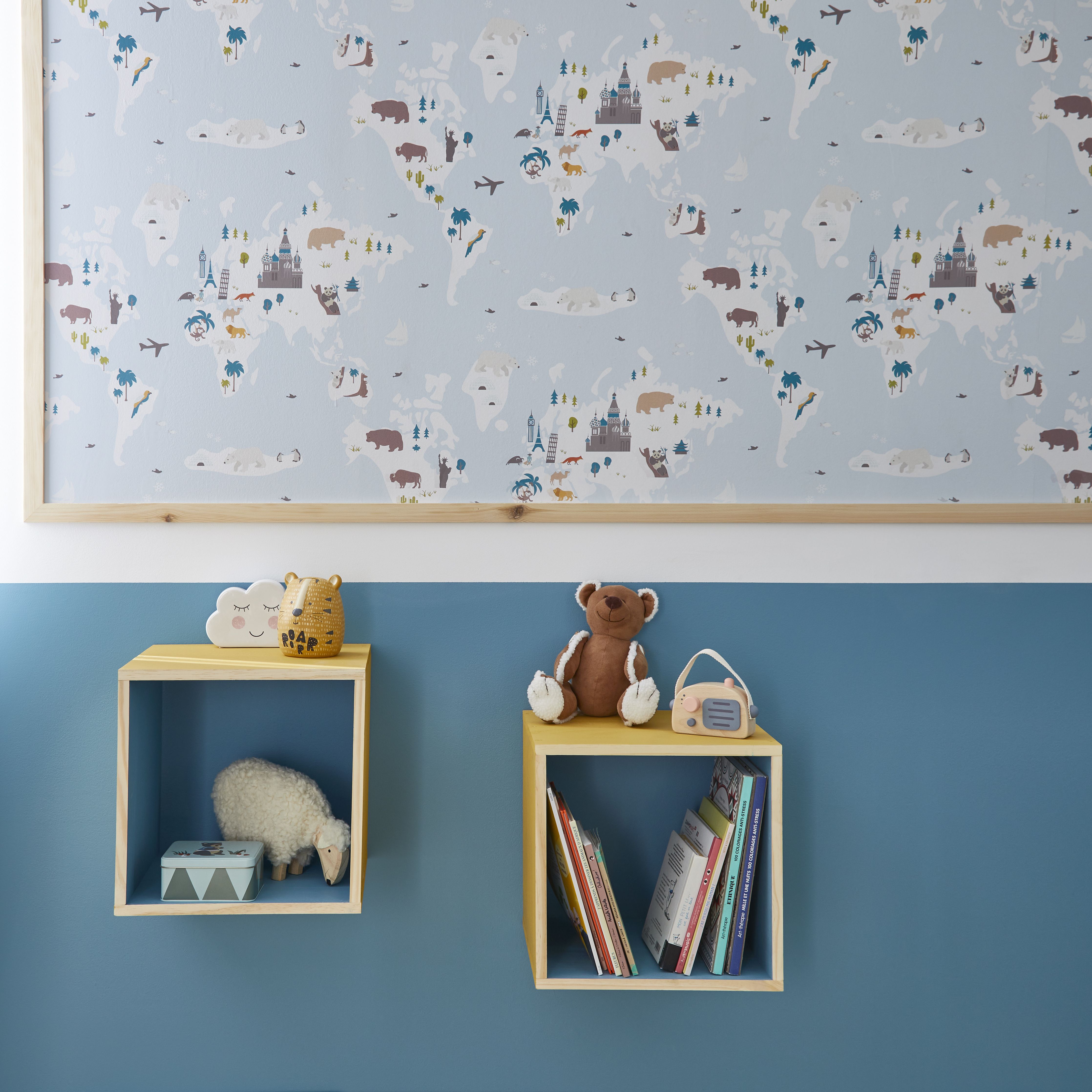 Children’s room featuring a blue and white world map wallpaper with illustrated animals and landmarks, above a teal painted lower wall. Wooden cube shelves display soft toys, books, and playful décor including a teddy bear and a small radio.