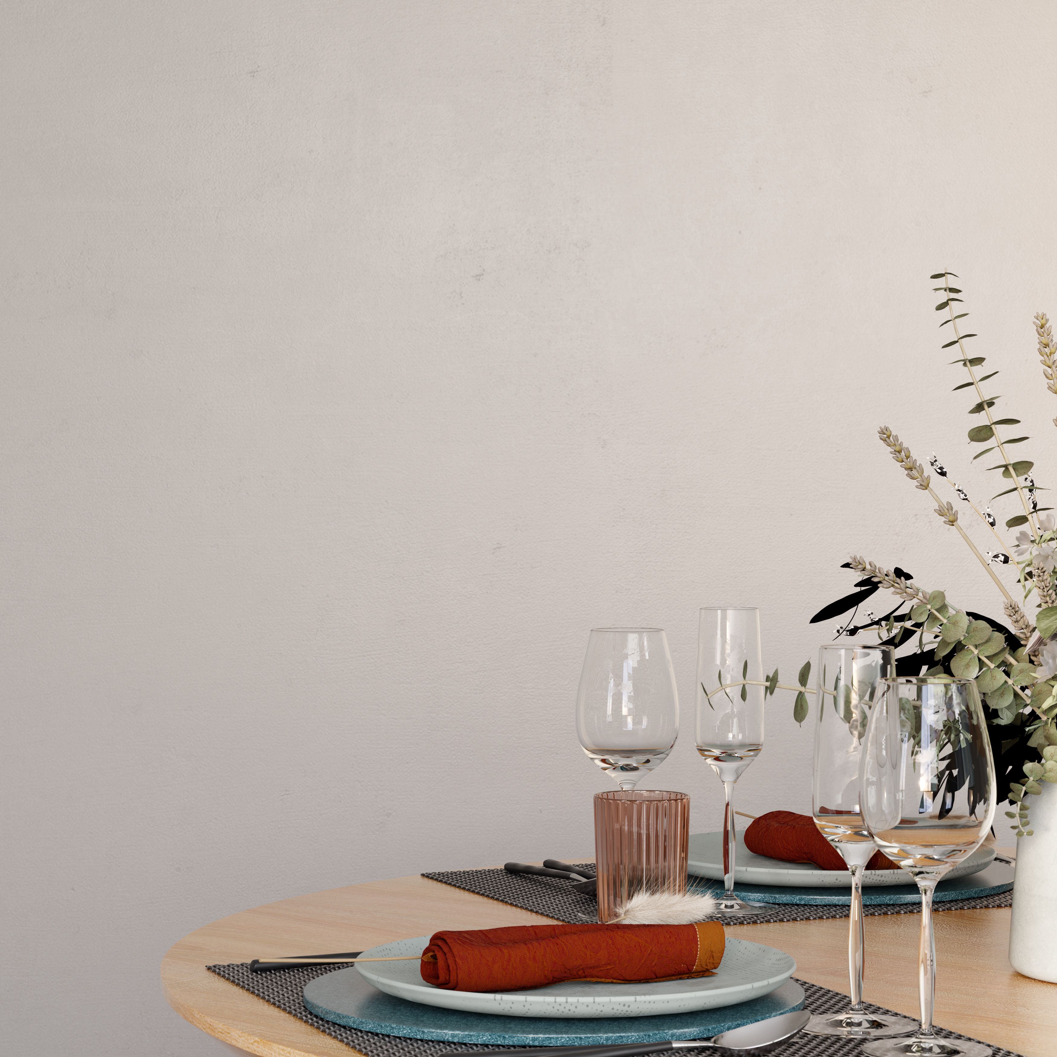 Dining table setting with GoodHome Tijuana painted wall in a soft warm neutral tone, featuring a round wooden table styled with plates, glassware, folded napkins, and a vase of mixed greenery and dried stems against a minimalist background with natural light casting soft shadows.