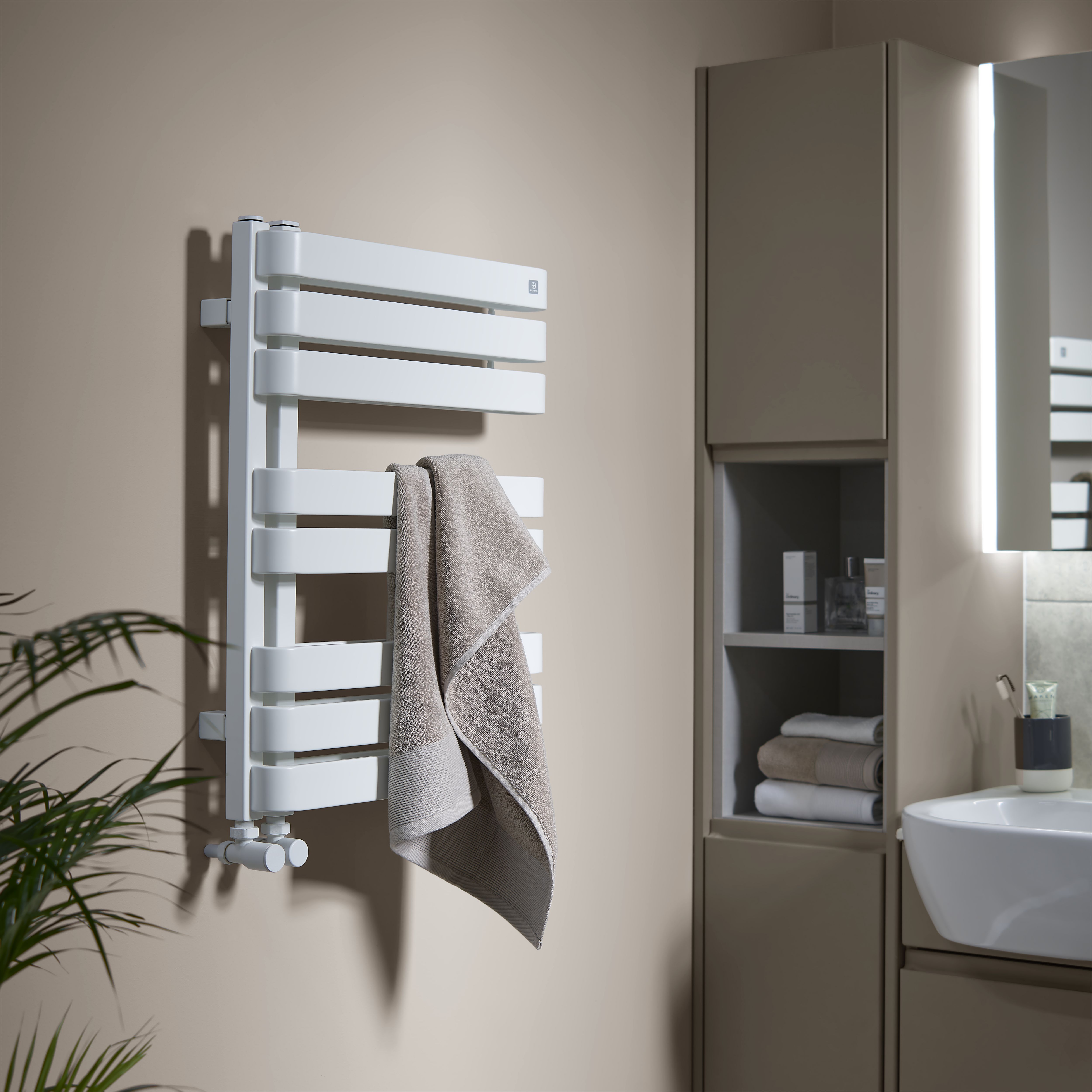 Bathroom with GoodHome Tijuana painted walls in a soft warm neutral tone, featuring a white heated towel radiator with a beige towel, a tall storage cabinet with open shelving, a sink unit with mirror, and soft lighting creating a calm, modern look.