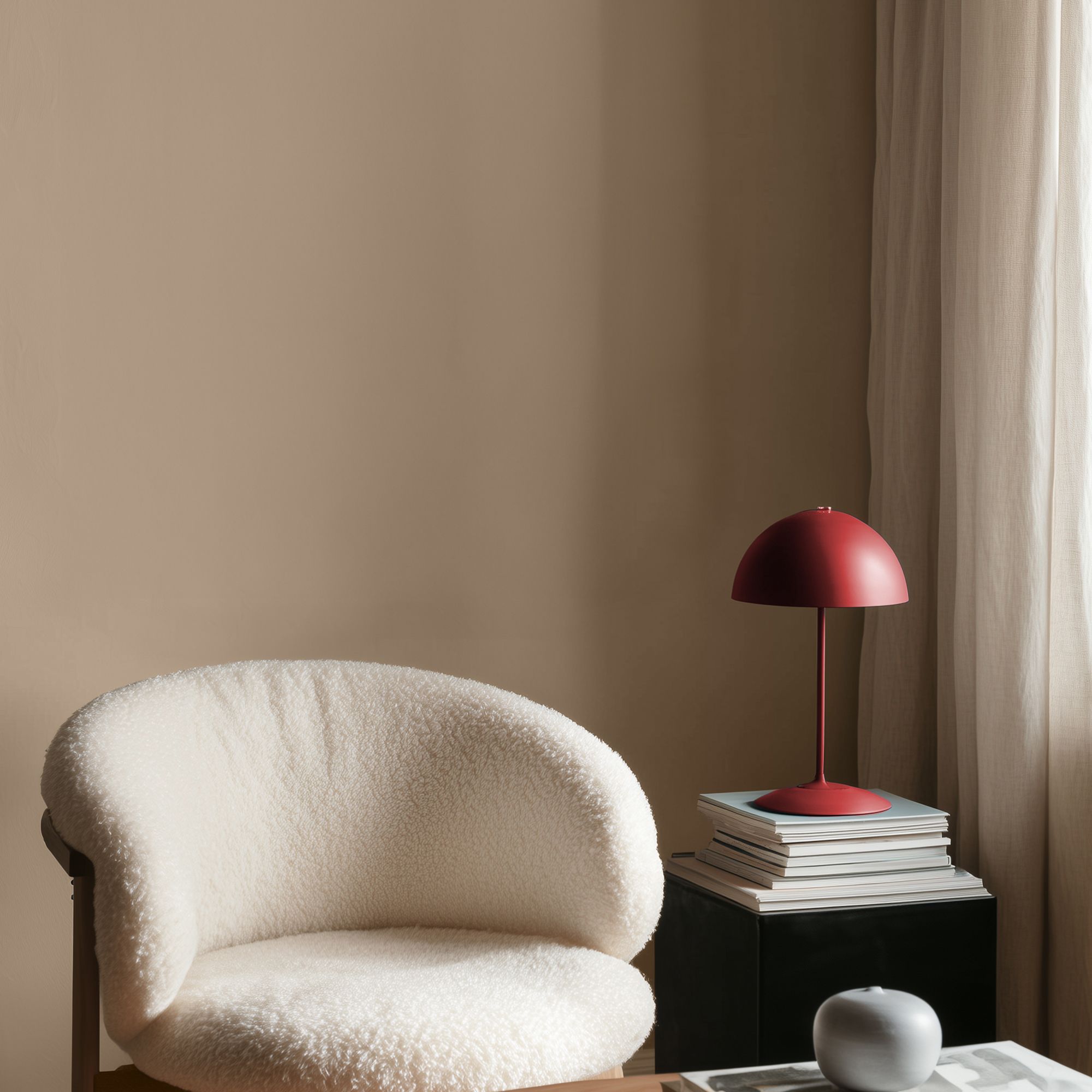 Living space with Lick Beige 02 painted walls in a warm neutral tone, featuring a textured cream armchair, a small side table stacked with books, and a red table lamp, with soft natural light filtering through sheer curtains.
