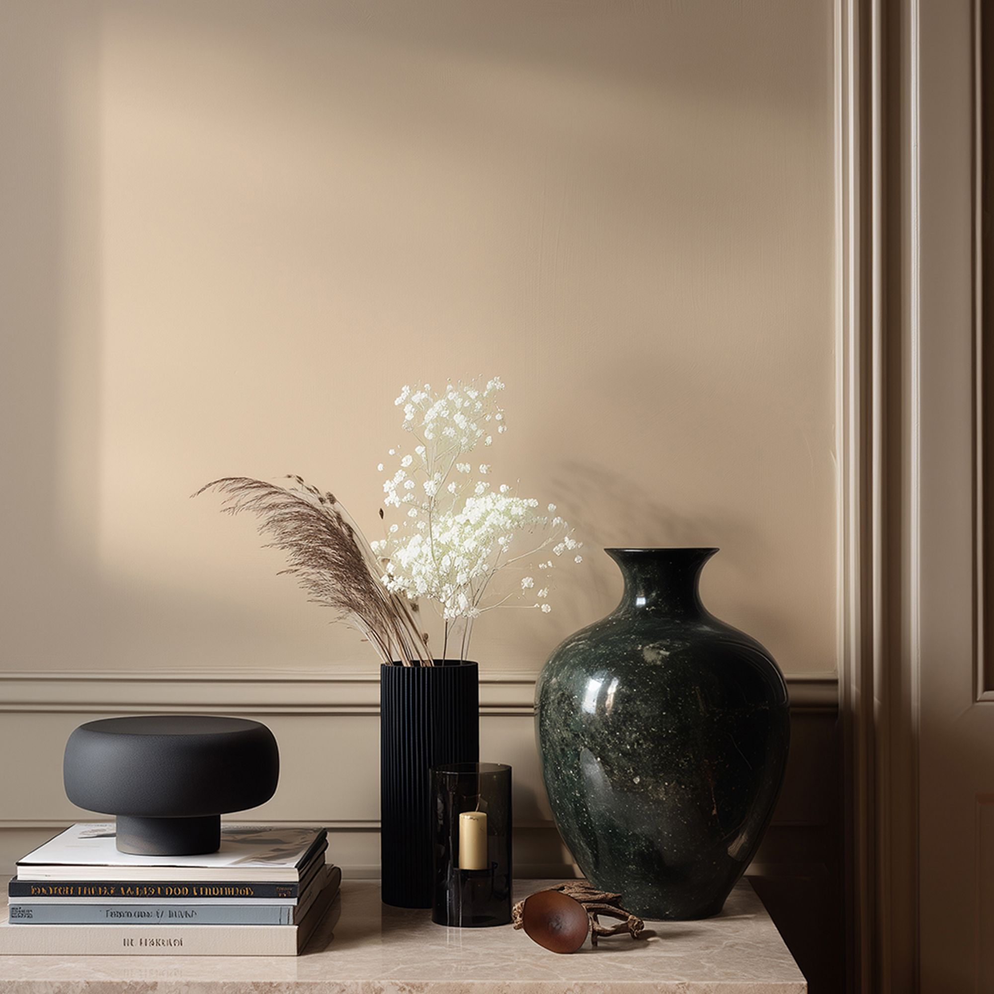 Interior vignette with Lick Beige 02 painted wall in a warm neutral tone, featuring a marble surface styled with stacked books, a black decorative bowl, a candle, and ceramic vases holding dried grasses and delicate white flowers, with soft natural light casting shadows.