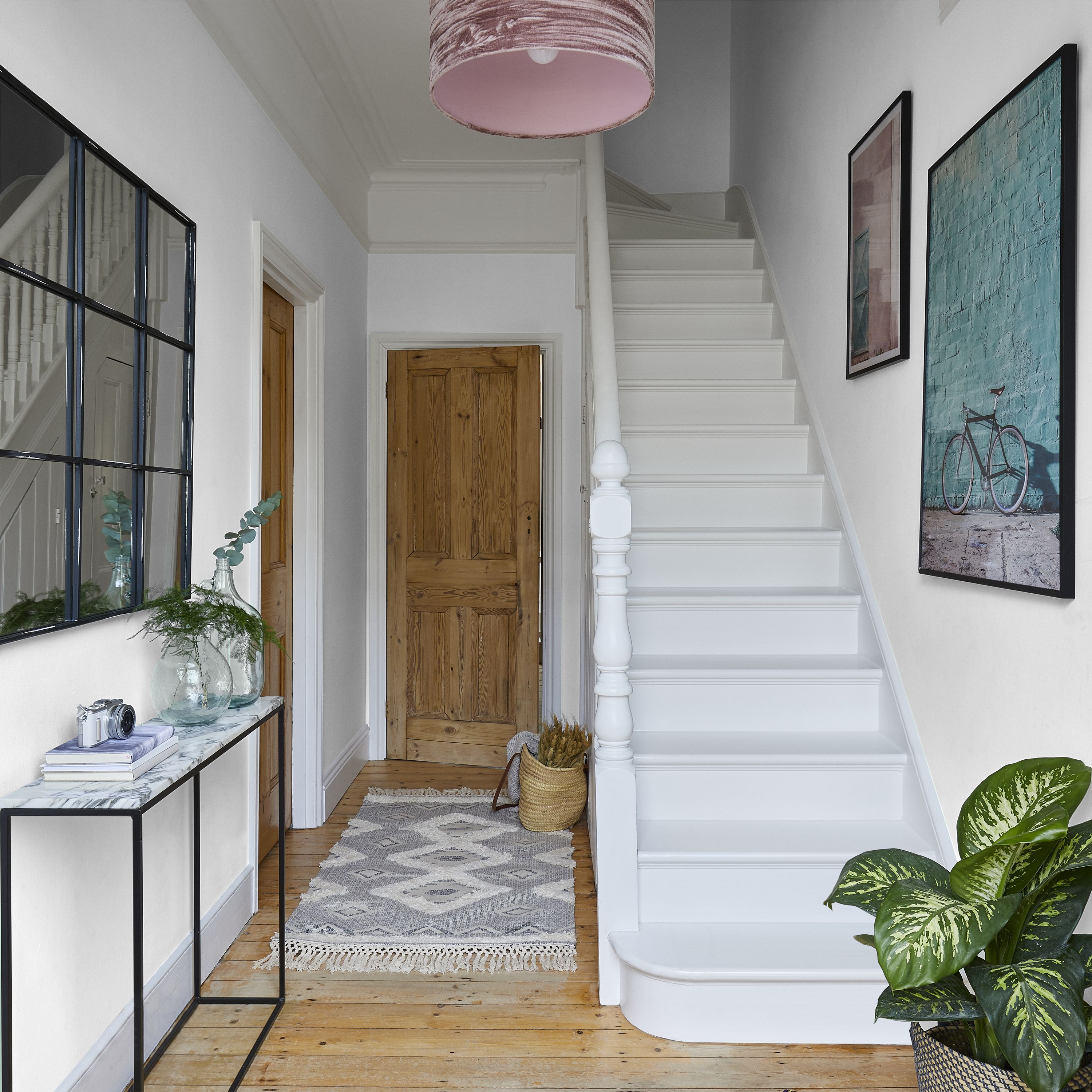 Hallway painted Valspar Rugged Olive, white staircase and banister, wooden doors, and light wood flooring. A patterned rug runs along the corridor, with a console table holding decorative items beneath a black-framed wall mirror. Framed artwork and a leafy houseplant add colour, while a pink ceiling lampshade sits above.