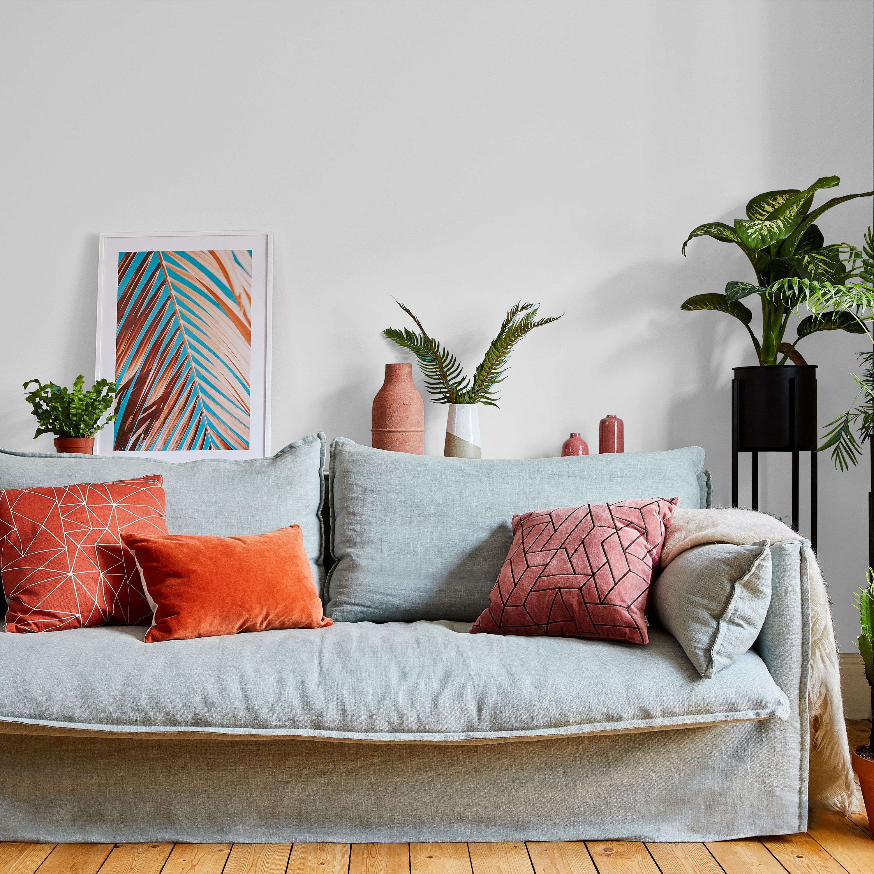 Living room with Valspar Rugged Olive painted walls, featuring a light grey sofa styled with coral and patterned cushions, wooden flooring, and indoor plants alongside framed artwork and decorative accessories.