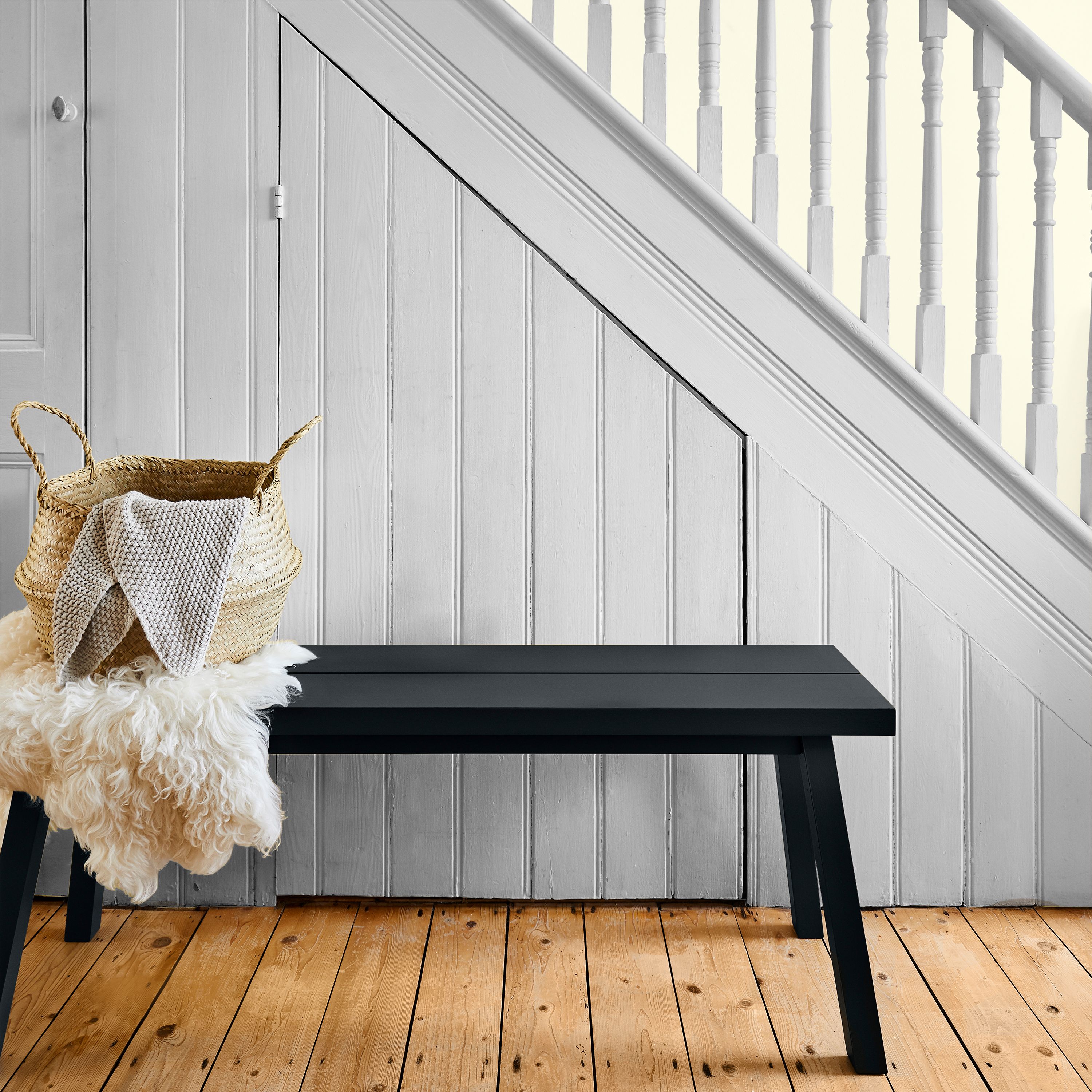 Hallway with Valspar Mellow Mango painted wood panelling in a warm peach tone, featuring a black bench, a woven basket with a throw, light wood flooring, and a white staircase creating a soft, inviting look.