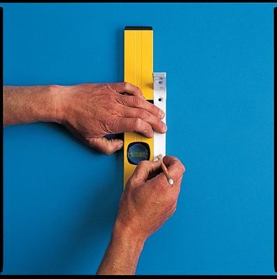 Person a holding a spirit level and shelf bracket on a wall, whilst marking the location of the screw holes on the shelf bracket in pencil.