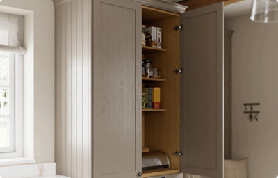 A wall-mounted shaker style kitchen cabinet in a warm grey colour is partially open, revealing glasses and boxed food. The inside of the cupboard is a warm, stained wood.