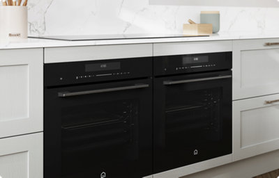 A dual-oven in black stands out against the light grey shaker style kitchen cabinets. Above, a black induction hob sits within a white and grey marble-effect countertop.