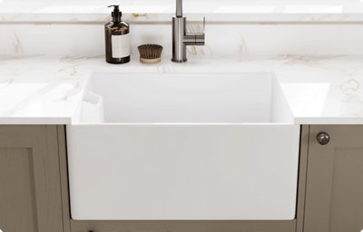 A white farmhouse kitchen sink matches is surrounded by a white marble-effect countertop and splashback. A brushed chrome kitchen tap glistens in the sunshine from the window behind.