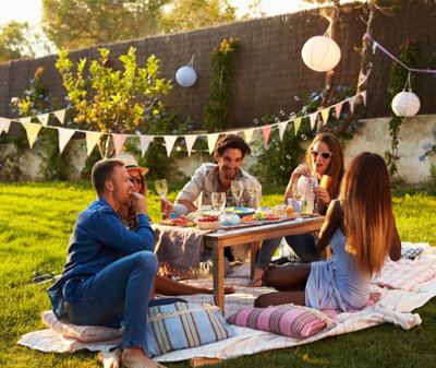 Group of friends sat around a wooden dining table on a blanket in a garden, surrounded by colourful bunting.