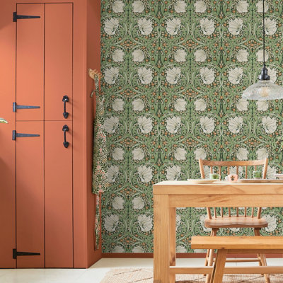 A dining room with classic green William Morris wallpaper featuring large white blooms and tiny orange flowers, complemented by a warm terracotta-painted cupboard.