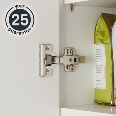 A close-up of a metal Kombu kitchen cabinet hinge on an open cupboard door. An icon on the image reads "25-year guarantee."
