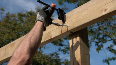 Person wearing gloves securing a horizontal timber beam to a vertical post using a coach bolt. Horizontal beam is resting on a nail fitted to the post and is clamped to the post at the top.