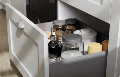 A deep shaker-style kitchen drawer in light grey is open to reveal various tall jarred foods. The drawer has a brushed chrome handle.