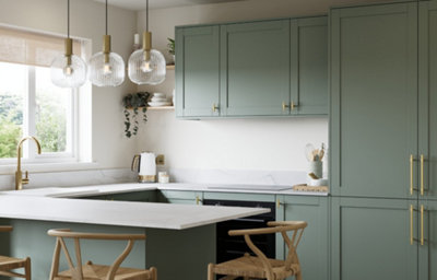 Reed green shaker kitchen with brass-effect handles, white marble-effect worktops and matching splashback. Features a central island with breakfast seating, tall integrated storage units, a Belfast-style sink with brass tap and decorative glass pendant lighting.