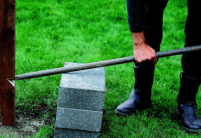 Demolition bar is being pushed into a notch cut out of a fence post. Person is resting the demolition bar on two breeze blocks to help lever the fence post out of the ground.