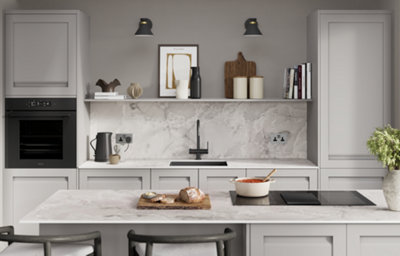 A woman is washing up at the kitchen sink while a man is holding a baby and looking happy. They are standing in a light grey U-shaped kitchen with white marble countertops.