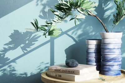A cosy bedroom corner with a side table holding a stack of books and a vase of flowers. Sunlight streams in, casting soft shadows on the blue wall, adding depth to the space.