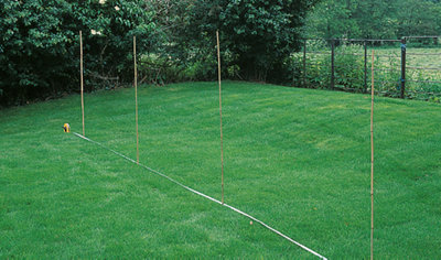 A garden lawn with four bamboo canes positioned in the ground and a tape measure stretched between them, marking out the planned location of a new fence.