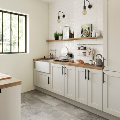 Kitchen with GoodHome Cancun painted walls in a soft warm neutral tone, featuring white cabinets with black handles, a farmhouse sink, wooden worktops, open shelving with plants and kitchenware, and natural light streaming through a window.