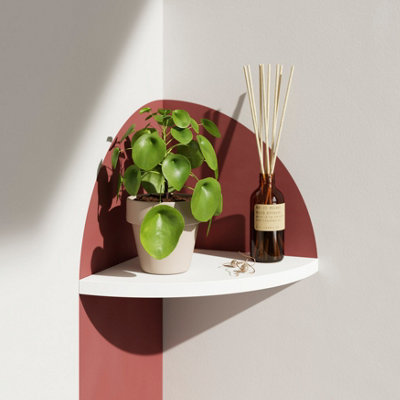 A floating white corner shelf in a white room with a potted plant and reed diffuser.