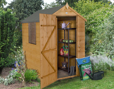 A small wooden shed with an apex roof in a garden surrounded by lush greenery.