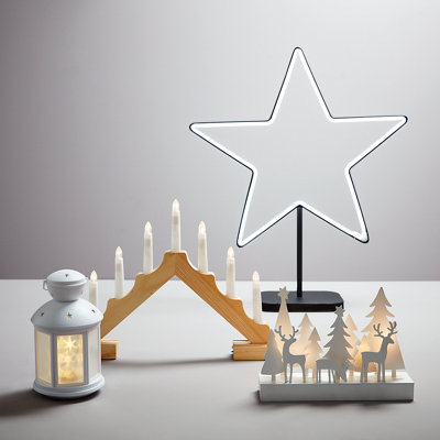 A selection of Christmas ornaments on a white table, including a black star light, wooden candelabra with artificial candles, small white lantern with cut-out stars, and a tree and deer silhouette ornament.