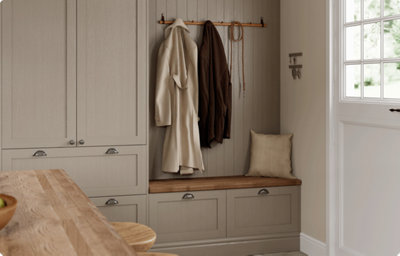 Floor to ceiling shaker kitchen cabinets are finished in a matt warm grey colour. There is bench seating in an alcove with matching shaker drawers below. The bench seat has a wood finish in a warm shade. Above, there are hook with coats hanging. Behind the coats, the wall is panels in the same matt warm grey finish to match the kitchen cabinets.
