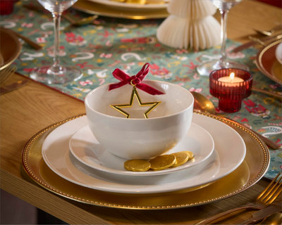 Close-up of two stacked white plates on a dining table, styled with a white napkin, rose gold cutlery, and a foliage and bauble decoration. Set on a white tablecloth with scattered baubles and glitter pinecones.
