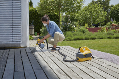 Person wearing a mask carefully applying paint to the surface of a garden deck using a paint sprayer.