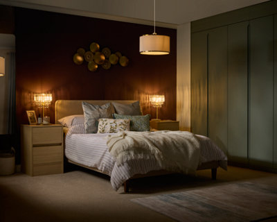 A modern bedroom with a double bed, striped bedding, throw blanket, and decorative pillows. Two wooden nightstands with lamps flank the bed. A sculptural wall piece and built-in green wardrobes are visible. A pendant light hangs overhead.