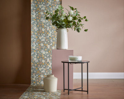 A beige vase with lush green leaves on a plinth, and a small round table with a white candle, are arranged in front of a floral wallpaper backdrop.