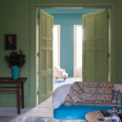 An interior with light green walls and open double doors in which there is a blue and white couch with a throw, a coffee table, a side table with a vase of flowers, and an armchair in the back.
