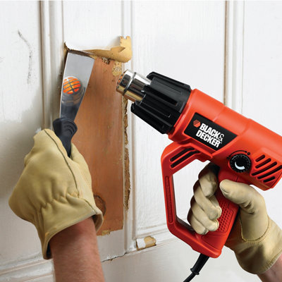 A close-up of a person's hands wearing gloves and removing white paint from wood using an orange heat gun and a scraper.