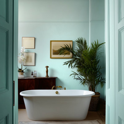 Bathroom with Lick Blue 09 painted walls in a soft teal blue, featuring a freestanding white bath, wooden cabinet with brass taps, framed artwork, and lush green plants creating a calm, spa-like setting.