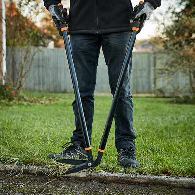 Person wearing gloves using grass shears to trim grass that has grown over the edge of the concrete edging around a lawn.