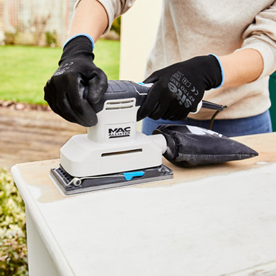 A person in a beige sweater removing white paint off of a wooden piece of furniture with a sanding tool while wearing black gloves.