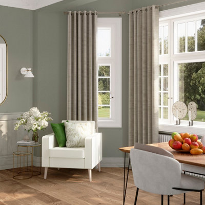 Bright living space with GoodHome Limerick painted walls in a soft green, featuring a white armchair with cushions, wooden flooring, large windows with neutral curtains, and a styled side table with flowers and decorative accessories.