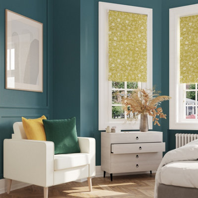 Room corner with GoodHome Saint Maxime painted walls in a rich teal blue, featuring a white armchair with green and yellow cushions, a light wood chest of drawers, a window with a patterned yellow blind, and a decorative vase with dried foliage.