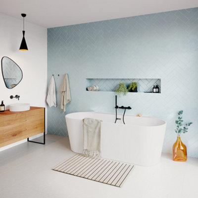 A double ended freestanding bath with black taps installed on a tiled wall behind the bath.