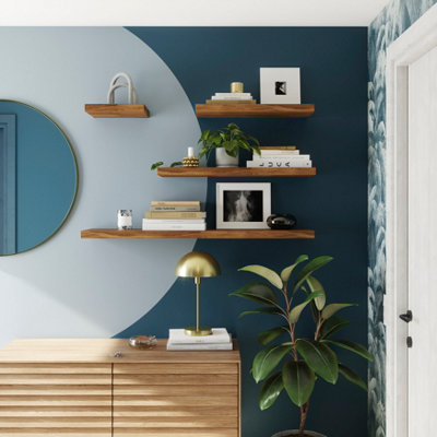 Four walnut effect wooden floating shelves attached to a wall, holding books, houseplants, candles and photo frames.