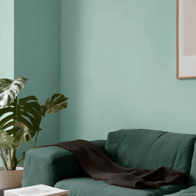 Living room with Lick Blue 09 painted walls in a soft teal blue, featuring a green fabric sofa with a draped throw, a large leafy houseplant, and minimalist wall art for a calm, modern look.