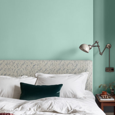 Bedroom with Lick Blue 09 painted walls in a soft teal blue, featuring a white upholstered headboard, layered bedding with cushions, a chrome adjustable wall lamp, and a wooden bedside table with decorative items.