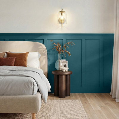 Bedroom corner with GoodHome Saint Maxime painted walls in a deep teal blue, featuring a white upholstered chair with green and mustard cushions, a light wood chest of drawers with a vase of dried flowers, and a window with a yellow patterned blind letting in natural light.
