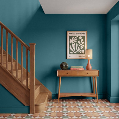 Hallway corner with GoodHome Saint Maxime painted walls in a deep teal blue, featuring a wooden staircase, a mid-century style console table with decorative objects, a table lamp, and framed abstract artwork above, set against patterned tiled flooring.
