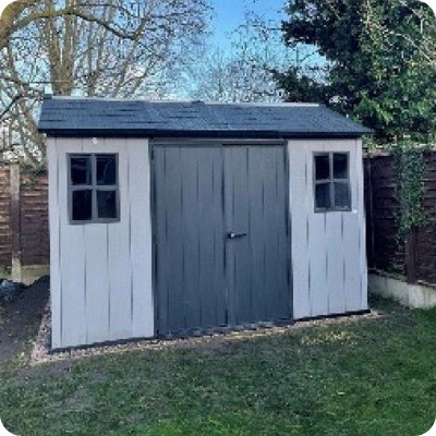 A light gray garden shed with black trim stands on a paved patio in a neatly kept backyard, next to a privacy screen and surrounded by lush greenery. A small seating area with outdoor chairs is set up beside the shed. This user-generated photo was shared from a real garden on Instagram with the handle @natandeuanshome