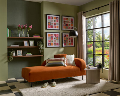 Living room with an orange chaise sofa against a green wall, styled with layered cushions, framed artwork, shelving with décor, a black floor lamp, large window with beige curtains, textured rug and small side table