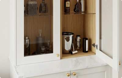A glass-fronted cupboard door is open to reveal cocktail making equipment and glass bottles. The interior of the cupboard is finished in a warm toned wood. The exterior has a matt finish and is a soft, creamy white colour.