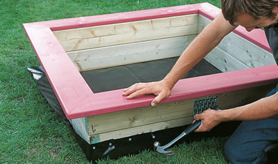 Person using a hammer and nails to secure weed control fabric to the bottom of a sandpit frame.