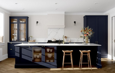 Elegant midnight blue shaker kitchen with a large island, marble-effect worktops and splashback, glazed display cabinets, brass hardware, wooden bar stools, and decorative floral arrangement.