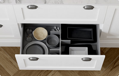 A shaker-style drawer is open to reveal a grey drawer interior with dividers in place to compartmentalise the contents. Various bowls and plates are inside. The drawer front is a soft, creamy white colour and has a matt finish. There are two antique looking, silver coloured handles on the drawer.